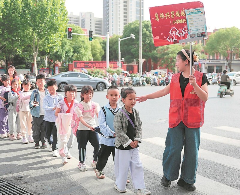 5月6日下午，合肥一六八玫瑰園學校南校區(qū)門口，身著紅色志愿者馬甲的“共享媽媽”丁艷和安徽大學社會學系志愿者劉詩琪手持“巢橋共享媽媽智習室”標牌，將十余名一年級學生護送至臨湖社區(qū)巢橋居民區(qū)的晚托課堂。.jpg